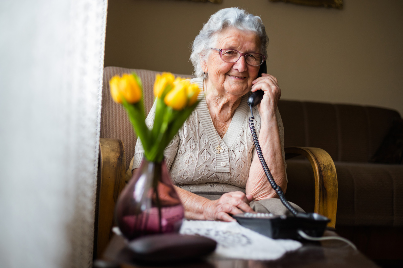 Elderly lady smiling whilst talking on the phone sitting on a chair in her living room.
